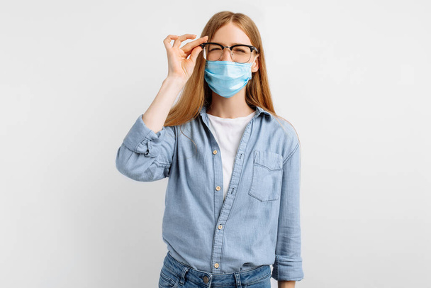 young woman in a medical protective mask on her face, wearing glasses for vision squints at the camera, having vision problems, on a white background - Фото, изображение
