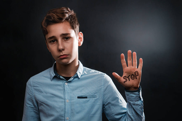 Het concept van huiselijk geweld. Een trieste tienerjongen laat een handpalm zien die zegt STOP. Begrepen, ruimte. Zwarte achtergrond. - Foto, afbeelding