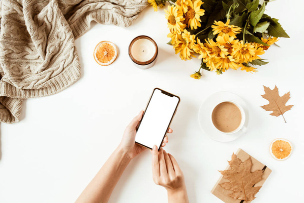 Women hand hold cell phone. Home office table desk workspace. Flat lay, top view girl boss business concept. Blanket, flowers bouquet, leaves, coffee cup. Template for social media. - 写真・画像