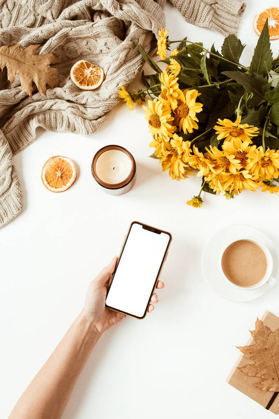 Women hand hold smartphone. Home office table desk workspace. Flat lay, top view girl boss business concept. Blanket, flowers bouquet, leaves, coffee cup. Template for social media. - Fotoğraf, Görsel
