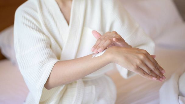 A happy beautiful woman in white bathrobe applying body moisturizing cream in bedroom, skin care and treatment concept - Photo, Image