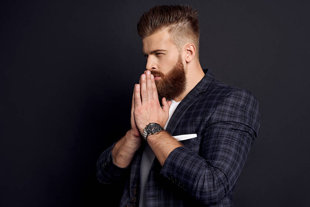 Thinking about something. Confident man with perfect hairdo and beard holds his hands to his face and prays, standing against grey background. - Photo, Image