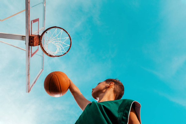 A young teenager in a green tracksuit is playing basketball, throwing a ball into the ring. Copy space. Bottom view. The sky in the background. Concept of sport and activity. - Valokuva, kuva