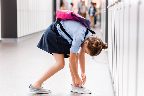 schoolgirl with backpack tying laces on gumshoe in school corridor - Photo, Image