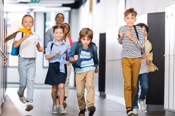 allievi multiculturali eccitati che corrono lungo il corridoio scolastico con insegnante sullo sfondo - Foto, immagini