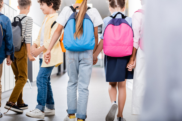 vista trasera de compañeros de clase multiétnicos tomados de la mano mientras caminan por el pasillo de la escuela, enfoque selectivo - Foto, Imagen