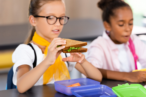 selective focus of schoolgirl in eyeglasses holding sandwich near african american classmate in school eatery - Photo, Image