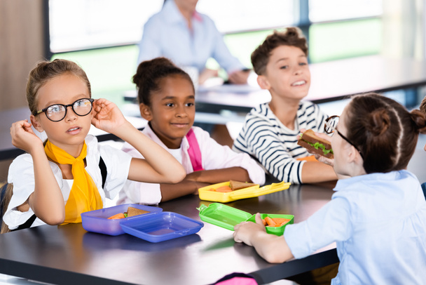 selective focus of multiethnic pupils sitting in school dining room near lunch boxes - Photo, Image