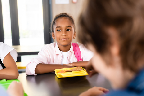 selective focus of african american schoolgirl sitting in school eatery near lunch box - Photo, Image