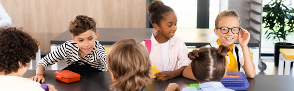 scolaretta che tocca gli occhiali mentre siede con compagni di classe multietnici nella mensa scolastica, immagine orizzontale - Foto, immagini