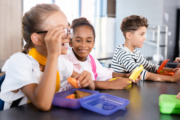 selektivní zaměření školačky dotýkání brýlí a držení čerstvé mrkve školní jídelně v blízkosti multikulturních přátel - Fotografie, Obrázek