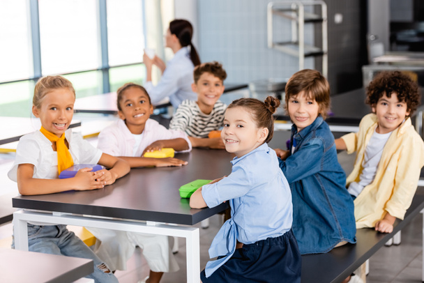 Multikulturelle Schüler blicken in die Kamera, während sie im Speisesaal sitzen, mit Lehrer im Hintergrund - Foto, Bild