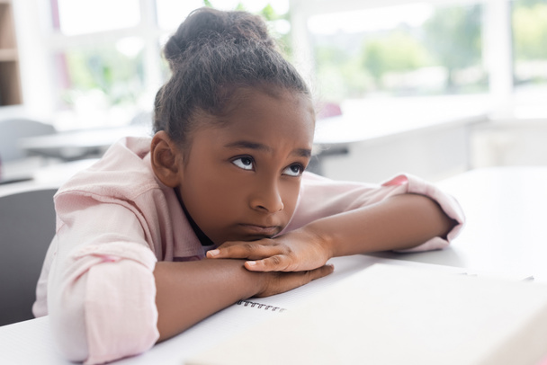 βαριέται αφροαμερικανή μαθήτρια κάθεται στο γραφείο με το κεφάλι στα χέρια κατά τη διάρκεια του μαθήματος - Φωτογραφία, εικόνα