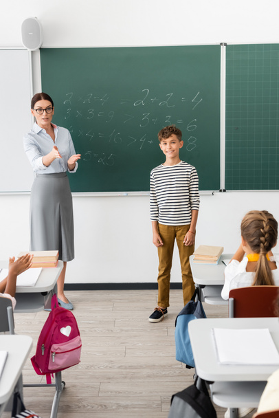 leraar wijzend met de hand terwijl staan in de buurt van schooljongen en schoolbord met vergelijkingen - Foto, afbeelding