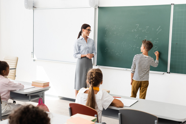 back view of schoolboy solving equations on chalkboard near teacher and multiethnic pupils - Фото, зображення