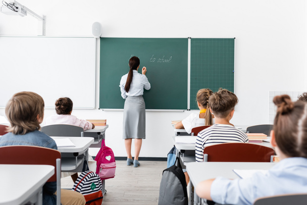 Rückansicht eines Lehrers in formeller Kleidung, der auf einer Tafel neben multiethnischen Schülern im Klassenzimmer steht - Foto, Bild
