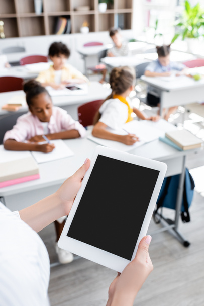 vista parziale dell'insegnante con tablet digitale con schermo vuoto vicino agli alunni multiculturali che scrivono durante la lezione - Foto, immagini