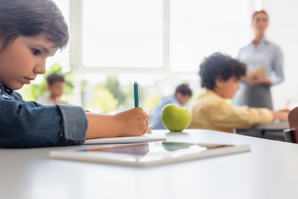 селективное внимание школьника письма во время урока рядом с яблоком и цифровой планшет - Фото, изображение