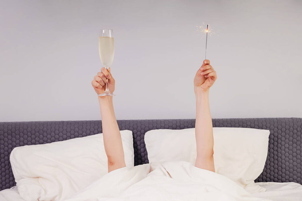 Hands holding glass of sparkling wine and sparkler while lying in bed. - Фото, изображение