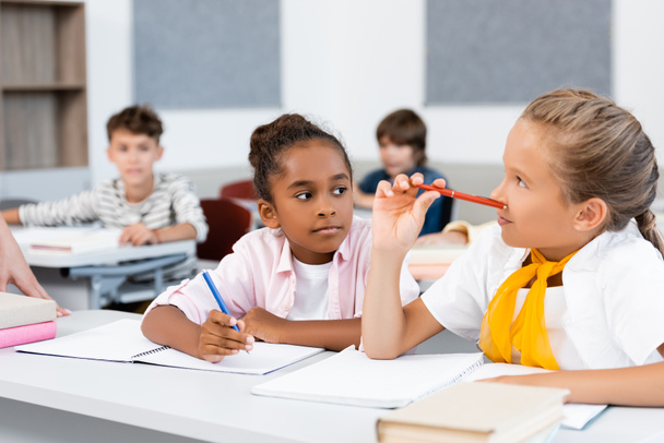 Selectieve focus van multi-etnische schoolmeisjes die op notitieboekjes schrijven tijdens de les in de klas  - Foto, afbeelding
