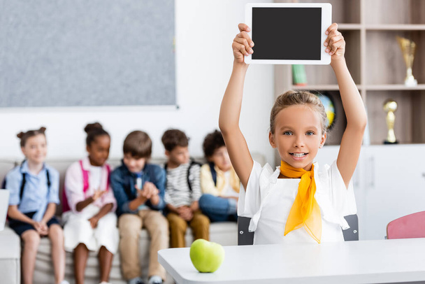 Selective focus of schoolgirl showing digital tablet near apple on desk and multiethnic classmates in classroom  - Foto, afbeelding