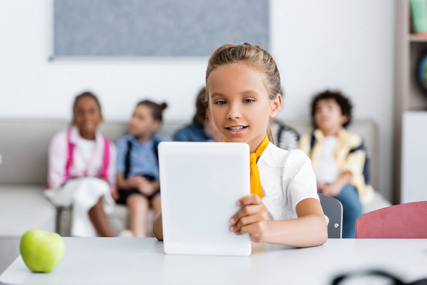 Selektivní zaměření školačky pomocí digitálního tabletu v blízkosti jablek a multikulturních přátel na pozadí ve třídě  - Fotografie, Obrázek