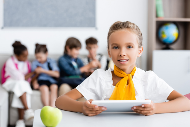 Selective focus of schoolgirl holding digital tablet near apple on desk and multiethnic friends in classroom  - 写真・画像