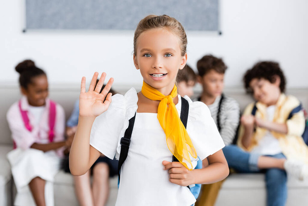Selective focus of schoolgirl with backpack waving hand at camera with multicultural friends at background  - Photo, Image
