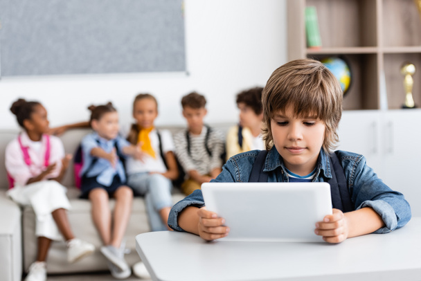 Selective focus of schoolboy holding digital tablet at desk with multicultural friends at background  - Zdjęcie, obraz