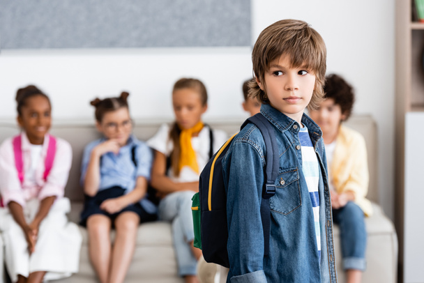Selective focus of schoolboy with backpack looking away near multiethnic friends in school  - Foto, afbeelding