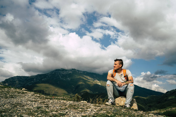 Young man sitting on hill. Dreamy male sitting on altitude and looking at Peaks of magnificent rocks located against bright cloudy sky. - Foto, immagini