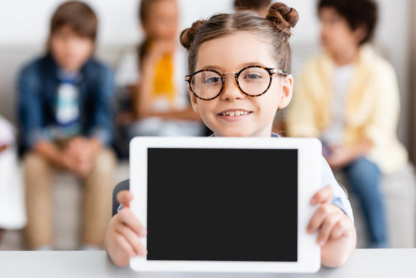Selective focus of schoolgirl in eyeglasses holding digital tablet at desk  - Foto, afbeelding