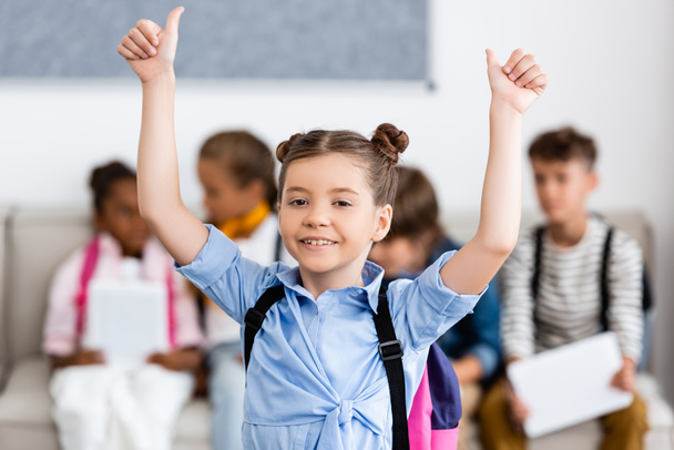 Selective focus o schoolgirl with backpack showing thumbs up near multiethnic friends at background  - Photo, Image