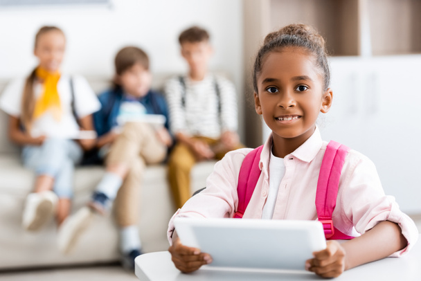 Enfoque selectivo de chica afroamericana sosteniendo tableta digital en el escritorio con compañeros de clase en el fondo en la escuela  - Foto, Imagen