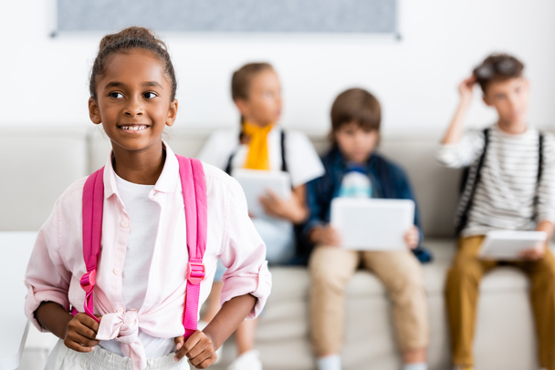 Szelektív fókusz afro-amerikai iskolás lány megható hátizsák barátaival a háttérben az osztályteremben  - Fotó, kép