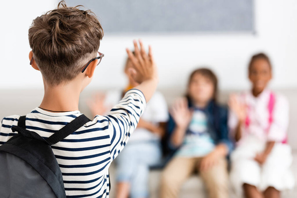 Selective focus of schoolboy with backpack waving hand at multiethnic classmates  - Foto, Bild