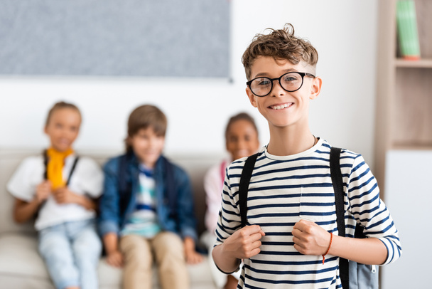 Вибірковий фокус школяра з рюкзаком і окулярами дивиться на камеру з однокласниками на фоні
  - Фото, зображення