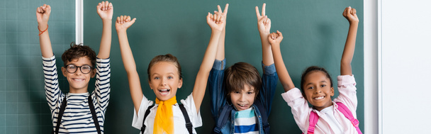 Horizontal crop of multiethic classmates with backpacks showing yeah and peace gestures near chalkboard  - Photo, Image