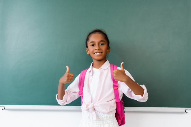 African american schoolgirl showing thumbs up near chalkboard in classroom  - Valokuva, kuva