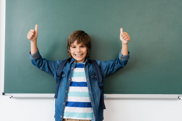 Schoolboy with backpack showing thumbs up near green chalkboard  - Fotoğraf, Görsel