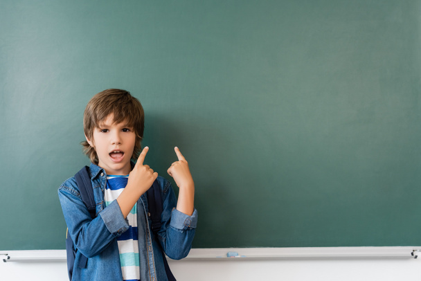 Écolier excité avec sac à dos pointant les doigts vers le tableau vert en classe  - Photo, image