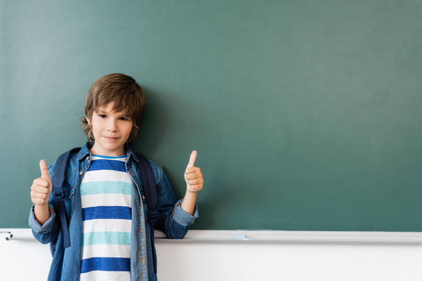 Iskolásfiú mutatja hüvelykujját, miközben áll a zöld tábla közelében  - Fotó, kép