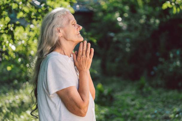 灰色の髪の祖母は屋外で祈っています。信仰、精神性、宗教の概念.  - 写真・画像