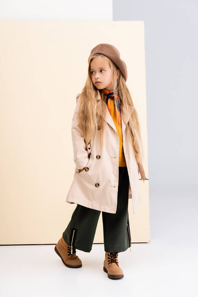 fashionable blonde girl in autumn outfit looking away and walking on beige and white background - Fotó, kép