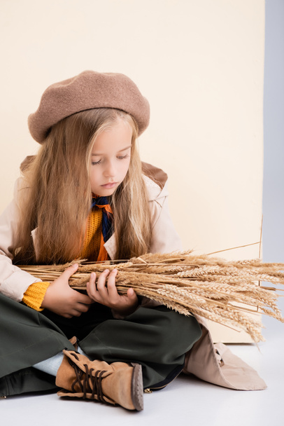 fashionable blonde girl in autumn outfit sitting on floor with wheat spikes on beige and white background - Photo, Image