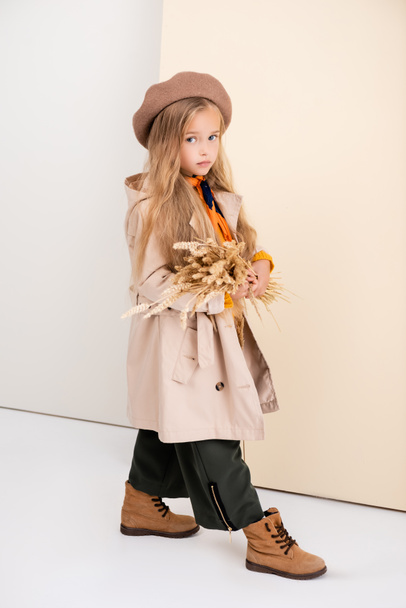 side view of fashionable blonde girl in autumn outfit walking with wheat spikes on beige and white background - Foto, immagini