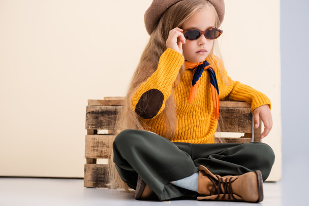 chica rubia de moda en traje de otoño y gafas de sol posando cerca de caja de madera sobre fondo beige y blanco - Foto, Imagen