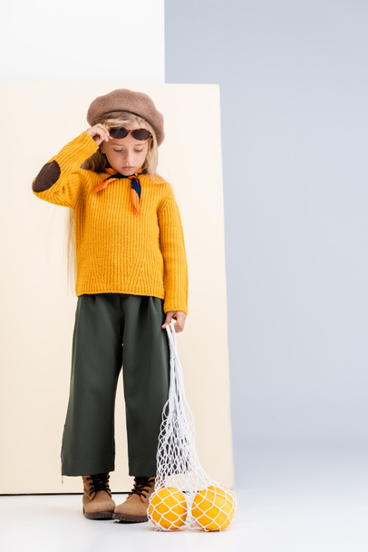 fashionable blonde girl in autumn outfit and sunglasses posing with grapefruits in string bag on beige and white background - Fotó, kép