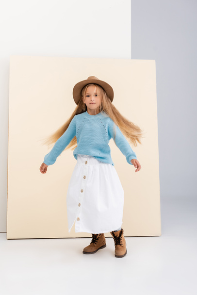 chica rubia de moda en sombrero marrón y botas, falda blanca y suéter azul girando cerca de la pared beige - Foto, Imagen