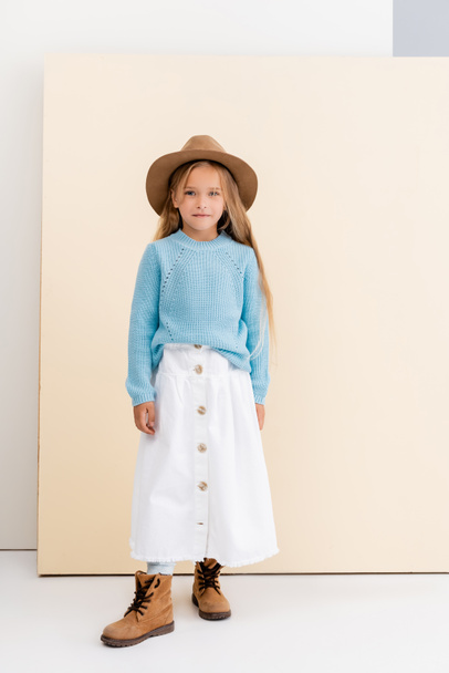 fashionable blonde girl in brown hat and boots, white skirt and blue sweater near beige wall - Photo, Image